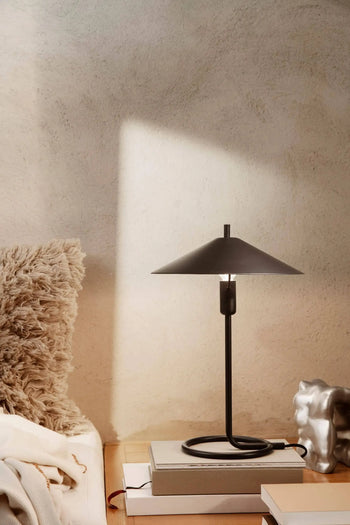 The ferm LIVING Filo Table Lamp, featuring a cone-shaped, powder-coated iron shade, sits on books beside a silver accent and textured beige pillow as sunlight casts geometric shadows—ideal for a modern home’s cream wall.