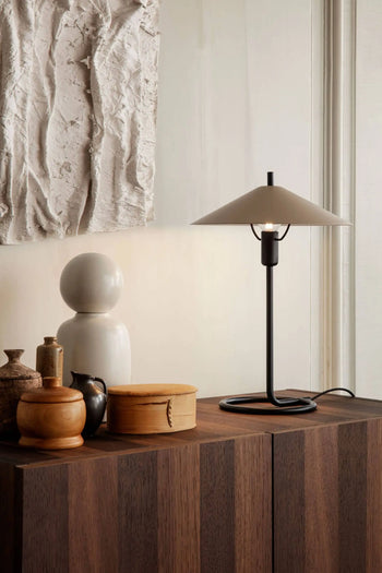 The ferm LIVING Filo Table Lamp, crafted from powder-coated iron with a white conical shade, rests on a dark wooden cabinet surrounded by decorative ceramics and wood accents—an elegant addition to any modern home.
