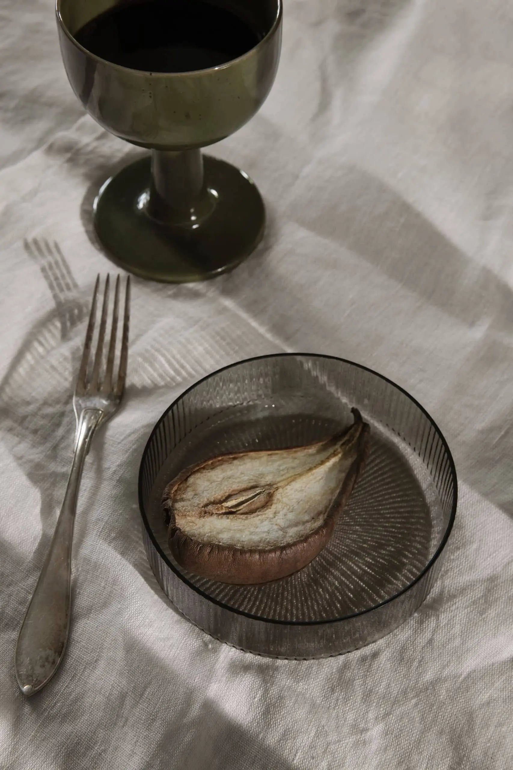 A glass of dark liquid in a ferm LIVING Floccula Wine Glass, a silver fork, and a sliced roasted pear on a ribbed plate rest on a cream tablecloth in soft light—capturing the ambiance of French bistros.