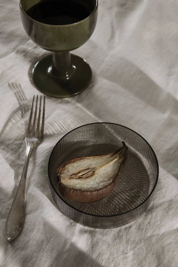 A glass of dark liquid in a ferm LIVING Floccula Wine Glass, a silver fork, and a sliced roasted pear on a ribbed plate rest on a cream tablecloth in soft light—capturing the ambiance of French bistros.