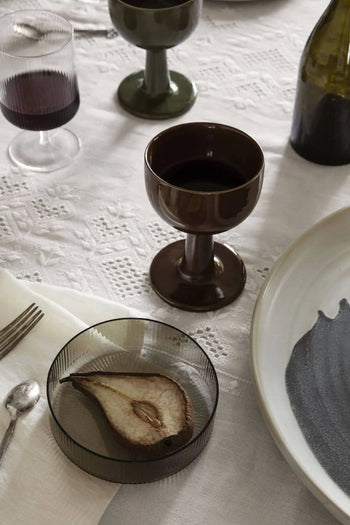 A table set with a ferm LIVING Floccula Wine Glass of red wine, a brown goblet, a plate with black detailing, a glass dish with dried pear, and cutlery on a white lace cloth evokes French bistro charm.