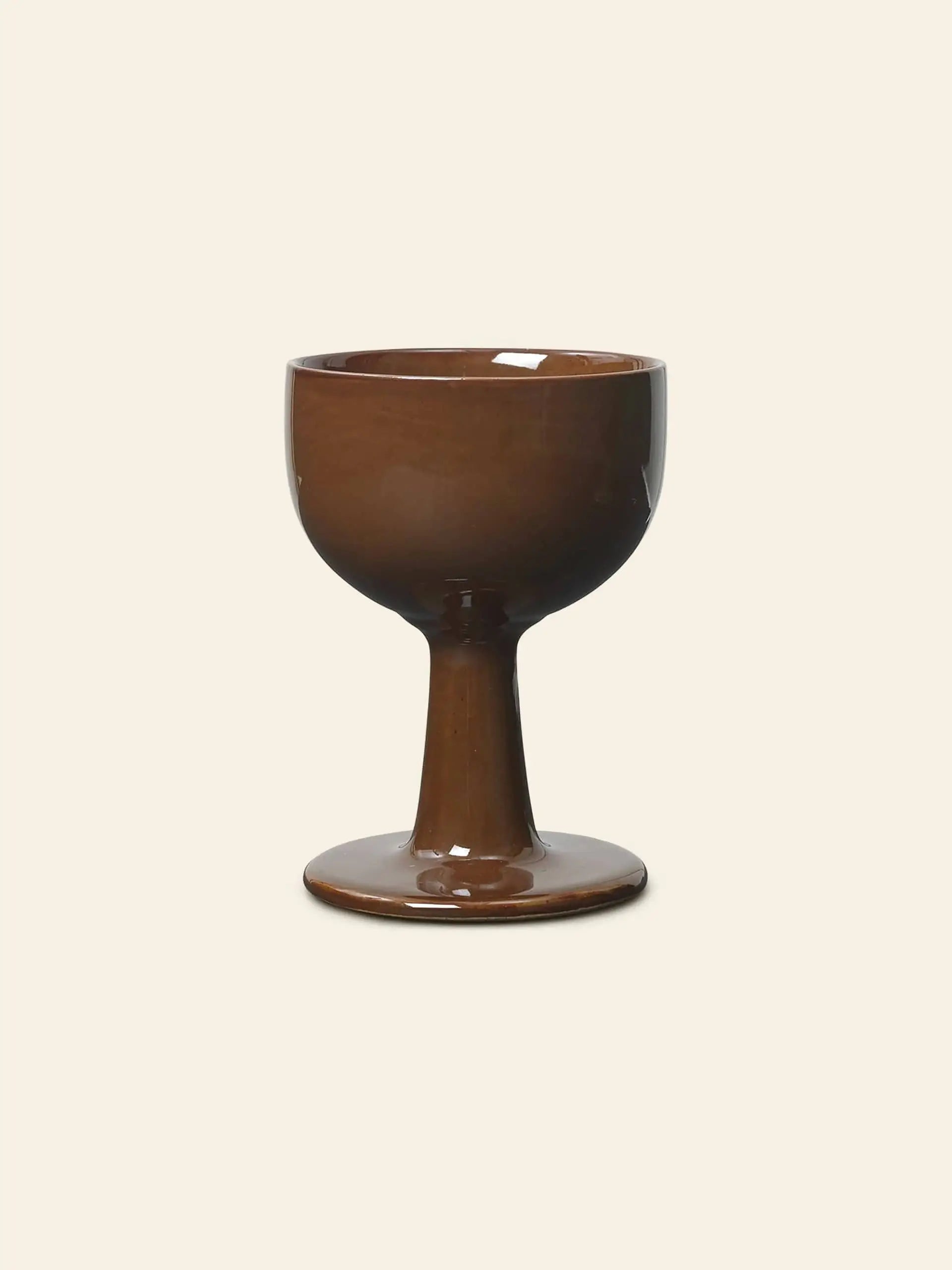 The ferm LIVING Floccula Wine Glass by ferm LIVING features brown ceramic with a wide, rounded bowl and sturdy, flared stem, echoing classic balloon wine glasses, shown against a plain beige background.
