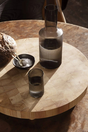 A ribbed glass carafe and matching glass by ferm LIVING stand on a round wooden table. Beside them, artisanal bread and a ferm LIVING Flow Porcelain Bowl, Small, with salt, sit on a wooden board in warm natural light.