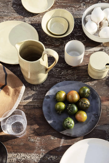 A rustic wooden table set with artisanal tableware, including ceramic dishes, a ferm LIVING Flow Porcelain Bowl, Small, a pitcher, cups, bowls, a gray plate with green tomatoes, garlic in a bowl, a napkin, and a clear plastic cup.