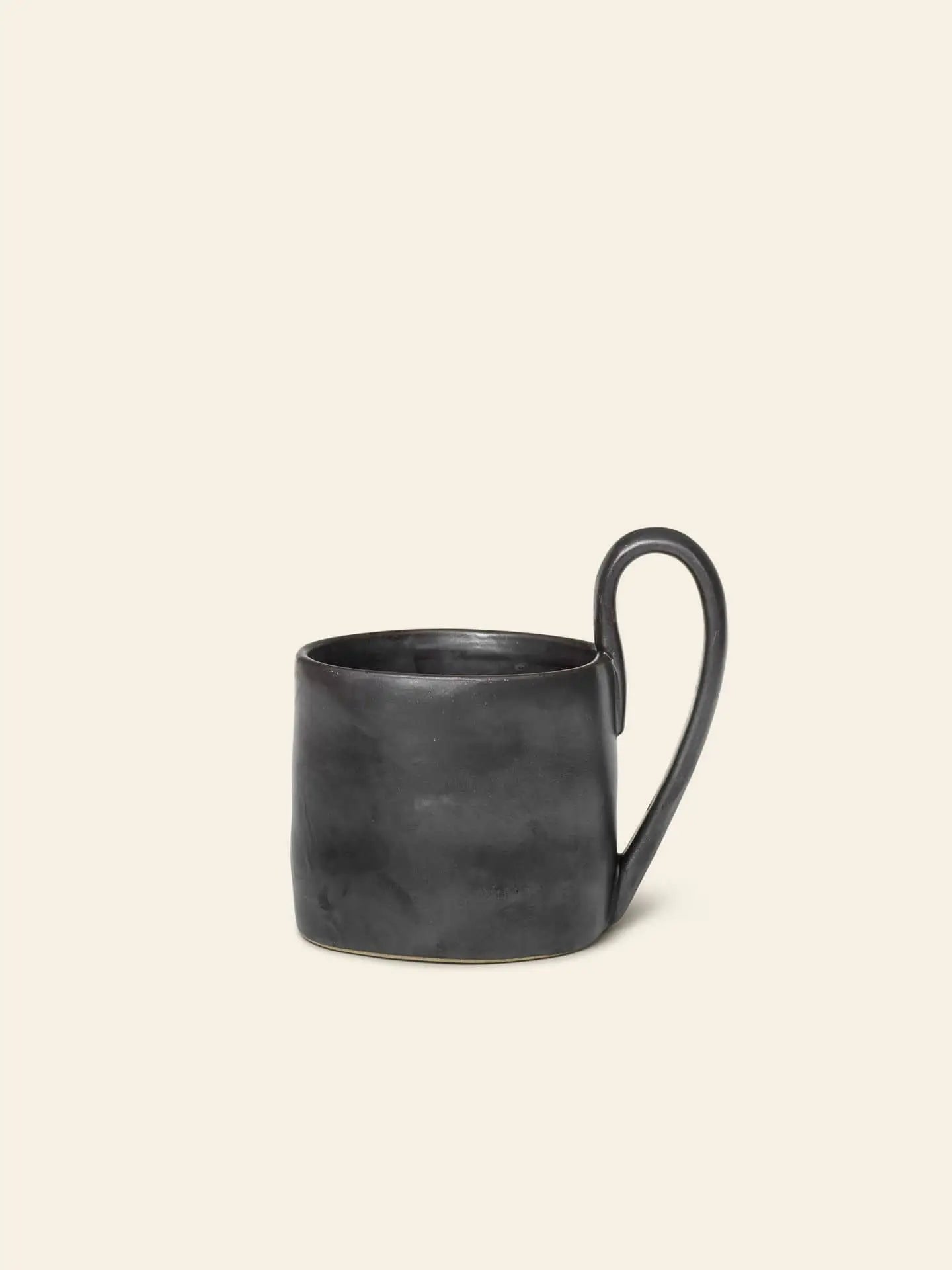 The ferm LIVING Flow Mug by ferm LIVING is matte black with an organic, slightly uneven surface and a distinctive elongated handle, shown against a plain cream background.