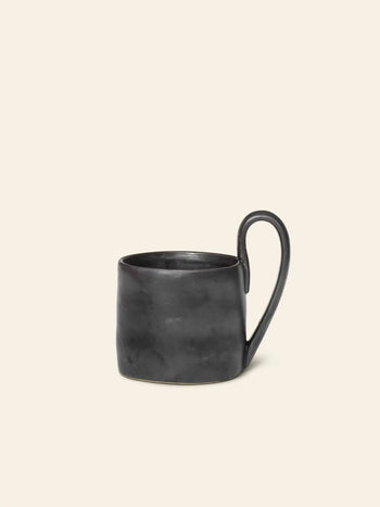 The ferm LIVING Flow Mug by ferm LIVING is matte black with an organic, slightly uneven surface and a distinctive elongated handle, shown against a plain cream background.