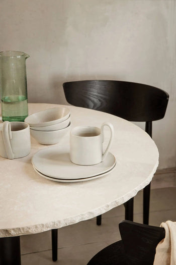 A round stone table with white ceramic plates, bowls, and the ferm LIVING Flow Mug by ferm LIVING sits near a glass pitcher and black wooden chair, adding to the minimalist aesthetic of the neutral room.