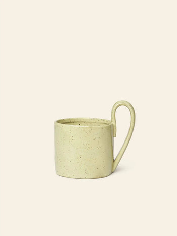 The ferm LIVING Flow Mug by ferm LIVING features a speckled light yellow finish, an organic design, and a large, uniquely shaped handle, shown on an off-white background for a minimalist aesthetic.