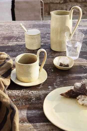 A rustic wooden table set with a yellow ferm LIVING Flow Mug, saucer, small jug, sugar bowl with spoon, glass of water, sugar cubes, and dark bread creates a warm scene highlighted by the organic design and minimalist striped cloth.