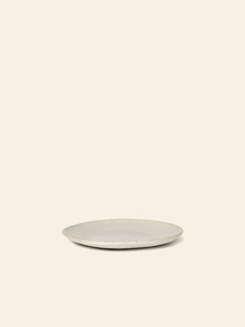 The ferm LIVING Flow Plate, Small, features a simple round porcelain design with a light reactive glaze, shown centered on a plain beige background with no other objects present.