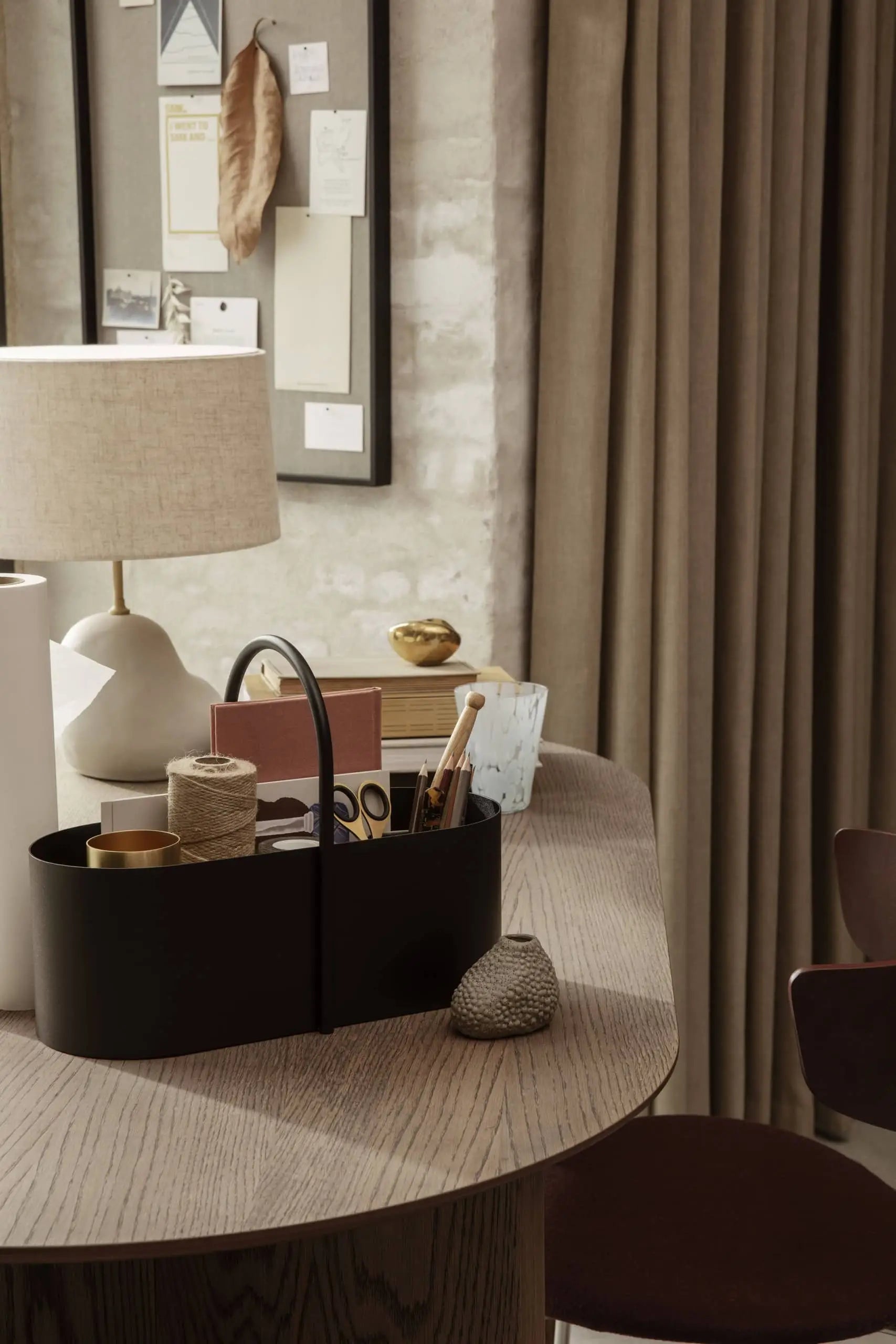 A cozy desk setup features the ferm LIVING Grib Toolbox by ferm LIVING, a slim organizer holding office supplies, paper rolls, twine, and brushes beside a beige lamp on wood. Neutral curtains and a pinboard with notes and photos are in the background.