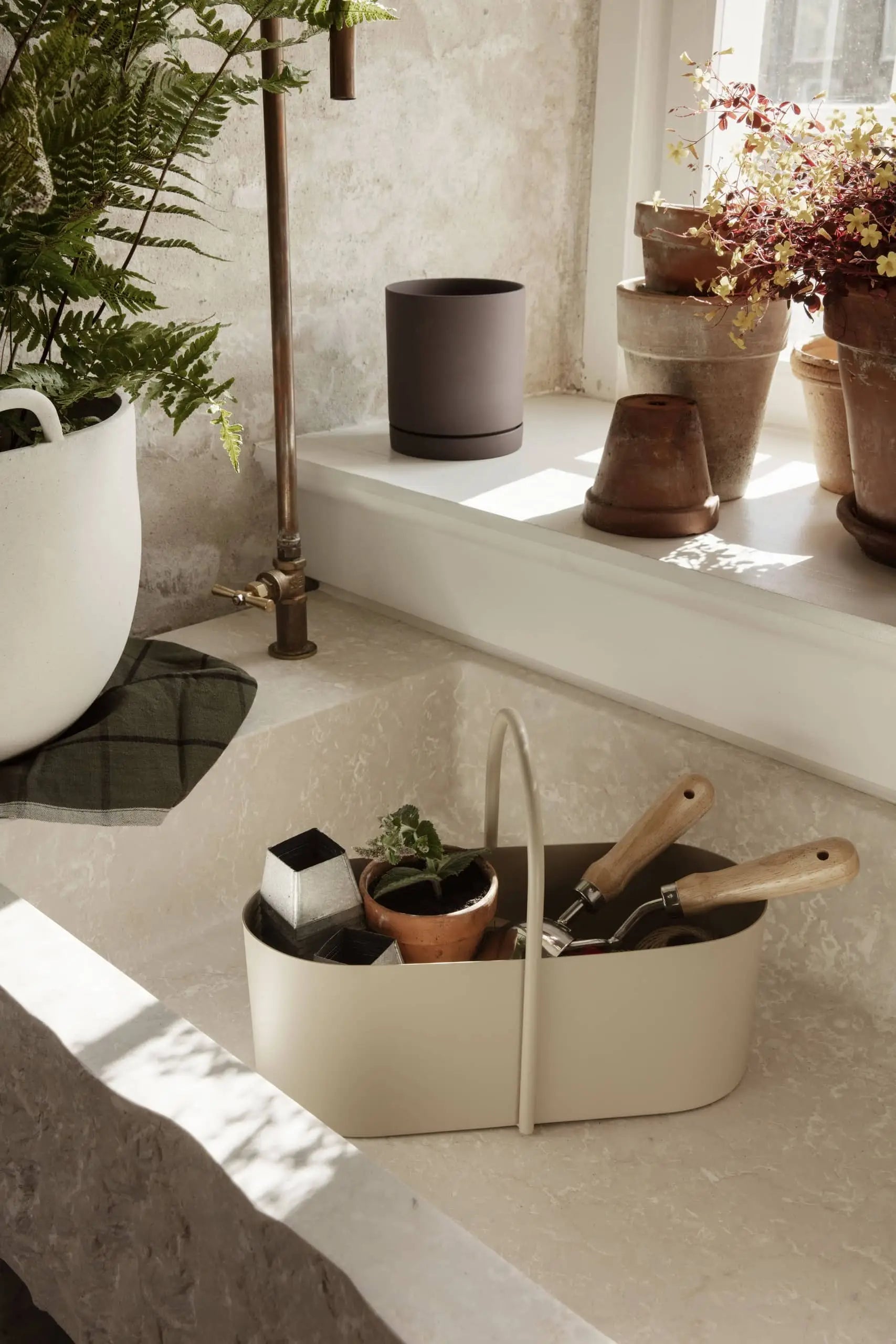 A cream-colored kitchen sink features the ferm LIVING Grib Toolbox by ferm LIVING as a caddy for gardening tools, alongside a potted plant and soap. Terracotta pots and plants fill the windowsill, with a green fern nearby. Sunlight brightens the space.