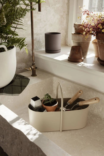 A cream-colored kitchen sink features the ferm LIVING Grib Toolbox by ferm LIVING as a caddy for gardening tools, alongside a potted plant and soap. Terracotta pots and plants fill the windowsill, with a green fern nearby. Sunlight brightens the space.