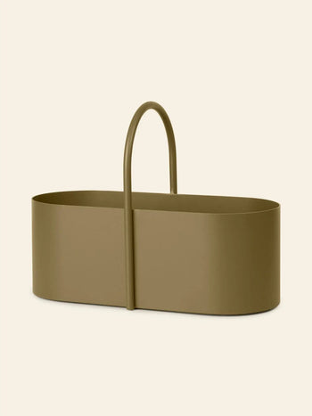 The ferm LIVING Grib Toolbox is an olive-green, oval metal organizer with tall sides, a slim silhouette, and a curved central handle, shown against a plain beige background.