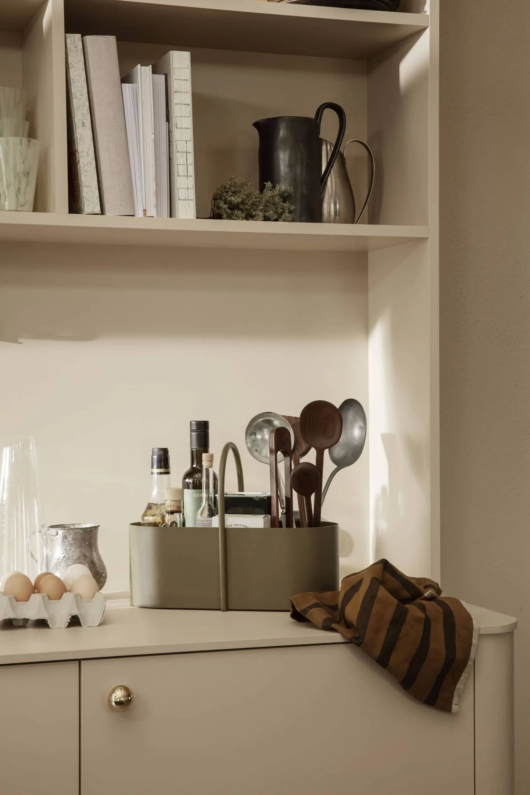 A kitchen counter displays a carton of eggs, clear glasses, a striped brown towel, and the slim ferm LIVING Grib Toolbox holding bottles and wooden utensils. Shelves above feature books, a metal pitcher, and decor.
