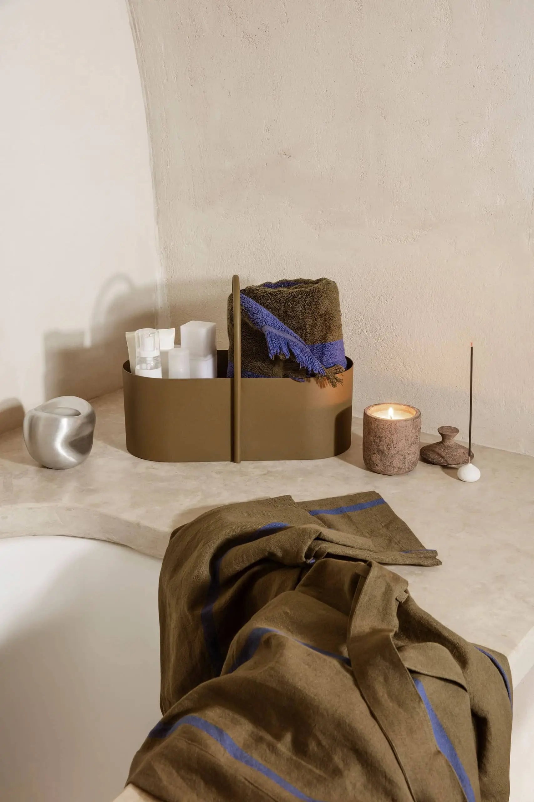 A minimalist bathroom scene showcasing the ferm LIVING Grib Toolbox in tan, holding toiletries and a rolled brown towel with a blue stripe, alongside a lit candle, incense holder, metallic cup, and matching towel draped over the tub.