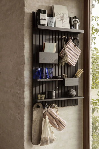 A modern black wall shelf displays books, blue glass cups, decor, pouches, the ferm LIVING Hale Oven Mitt (recycled polyester), cutting boards, and mesh bags beside a window with greenery visible outside.