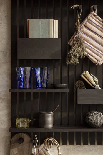 A dark wooden kitchen shelf with glassware, books, ferm LIVING Hale Pot Holders (set of 2), dried herbs, utensils, a small metal pot, a glass cup, and a dark textured object stands out against a light wall.