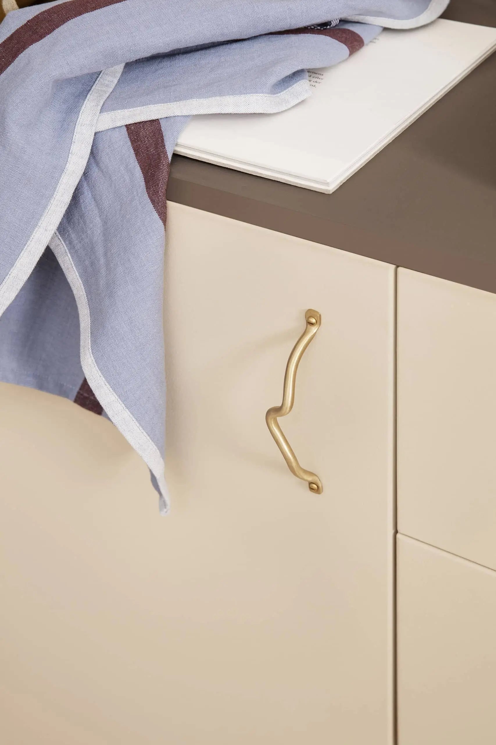 A beige kitchen cabinet with a gold wavy handle and brown countertop features the ferm LIVING Hale Tea Towel by ferm LIVING draped next to a white cutting board.