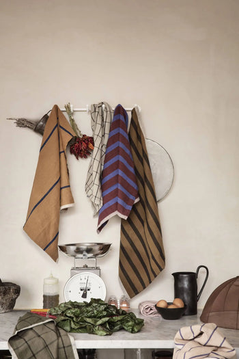 A kitchen scene featuring ferm LIVING Hale Tea Towels by ferm LIVING hanging on a rack, with dried flowers, a vintage scale, black pitcher, eggs, bottle, leafy greens, and a checkered cloth on a marble countertop.