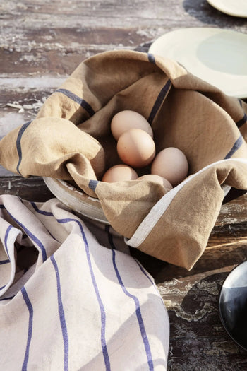 A rustic wooden table displays a cloth-lined bowl of brown eggs beside the ferm LIVING Hale Tea Towel and glimpses of plates.