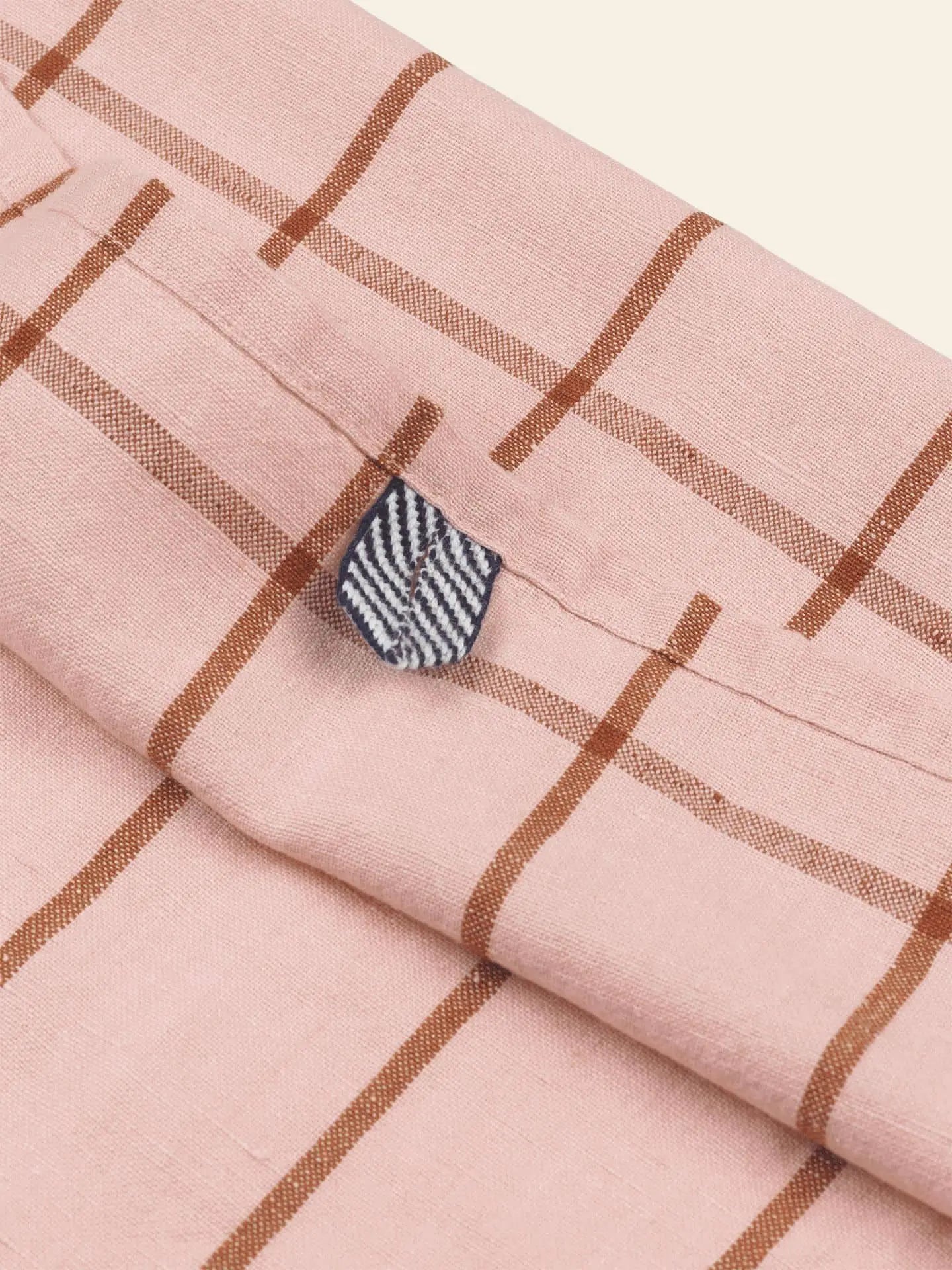 A close-up of the ferm LIVING Hale Tea Towel shows pink fabric with brown checks and a small navy and white striped tab sewn into the seam.