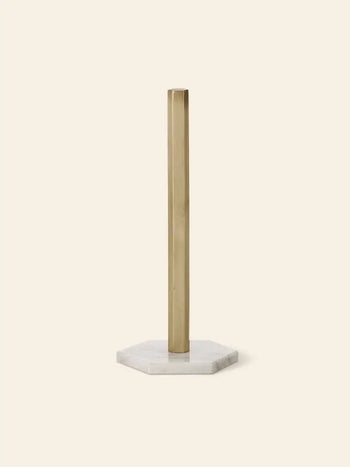 The ferm LIVING Hexagon Stand is a minimalist paper towel holder with a brass-finished vertical rod and white marble hexagon base, designed by ferm LIVING to hold rolls neatly on a light beige background.