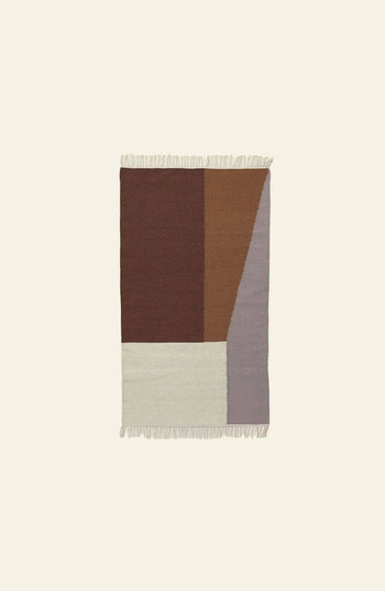 The ferm LIVING Kelim Rug is rectangular with an abstract geometric pattern in brown, tan, cream, and gray, plus fringe on the top and bottom edges. Its bold shapes create visual interest against a plain light background.