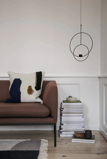 A modern living room featuring a brown sofa, the ferm LIVING Loop Cushion by ferm LIVING crafted from New Zealand wool, a stack of books with a green dish and cup on top, and a minimalist black geometric wall sculpture.