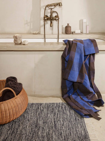 A modern bathroom with a large bathtub, luxury vintage-style faucet, blue and brown checkered towel over the edge, a wicker basket with dark towels, and the ferm LIVING Melange Rug by ferm LIVING on the floor.