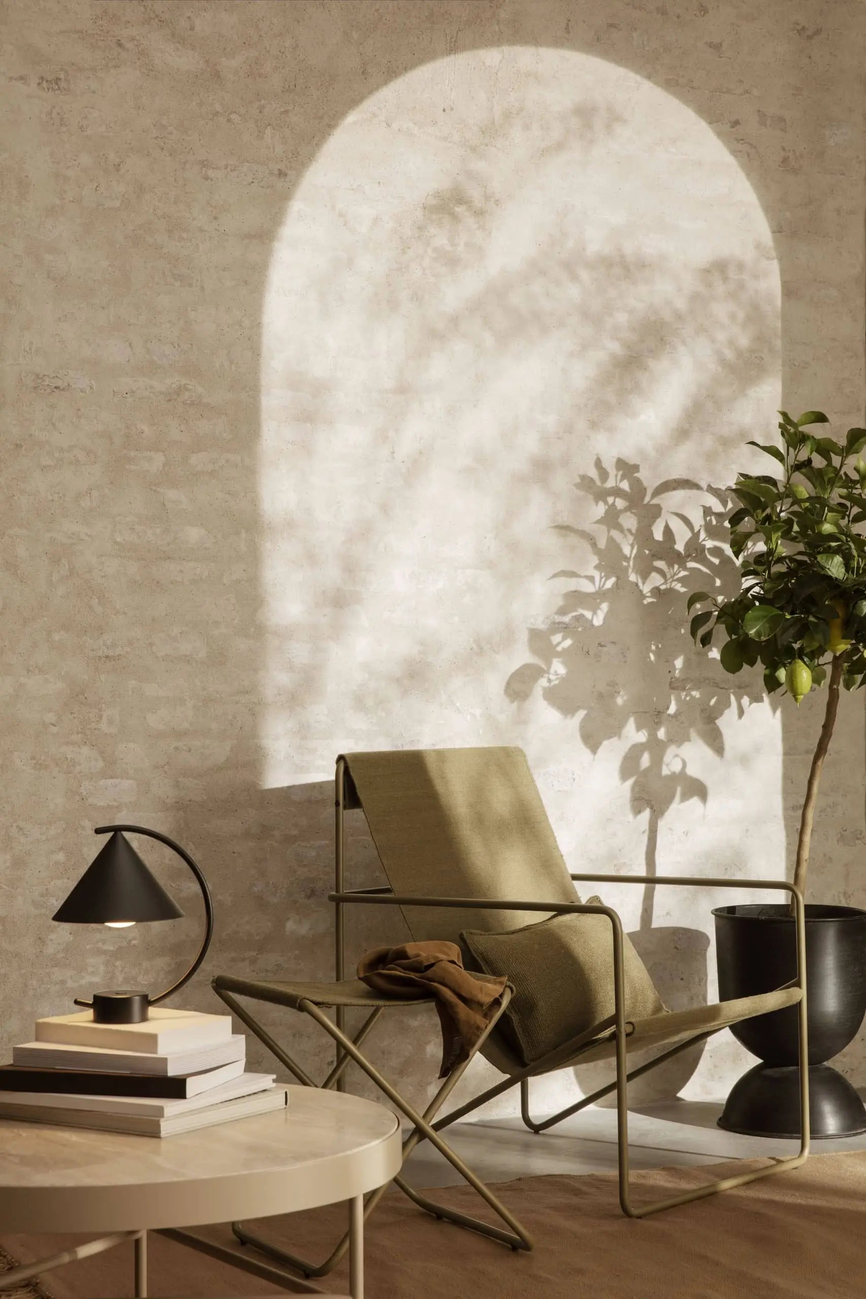 A cozy reading nook features a green lounge chair, a potted plant, and a round table with books. The sculptural ferm LIVING Meridian Lamp by ferm LIVING adds style as natural light casts arched shadows on the textured wall.