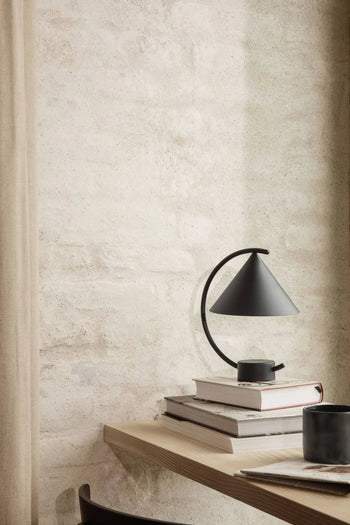 A minimalist workspace features a light wooden desk, stacked books, black cup, and soft beige walls. The ferm LIVING Meridian Lamp in powder coated steel softly illuminates the space, creating a calm and cozy atmosphere.