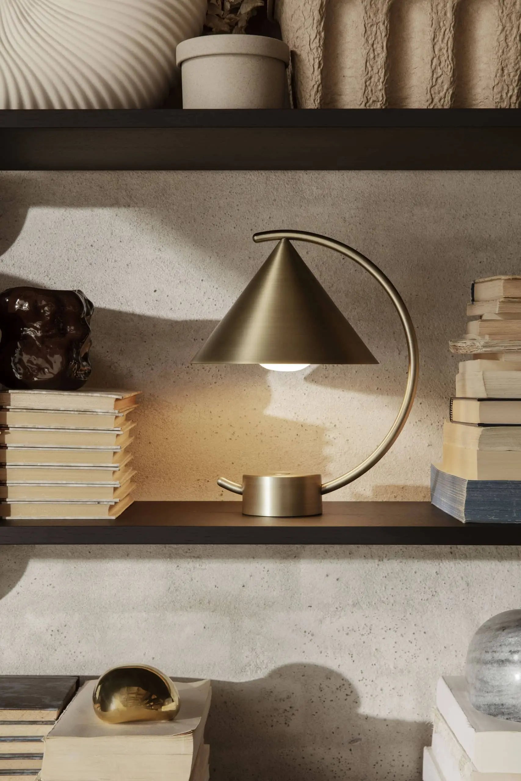 The ferm LIVING Meridian Lamp, featuring a sculptural brass design and conical shade, rests on a dark wooden shelf with stacked books and decor, all set against a textured light-colored wall.