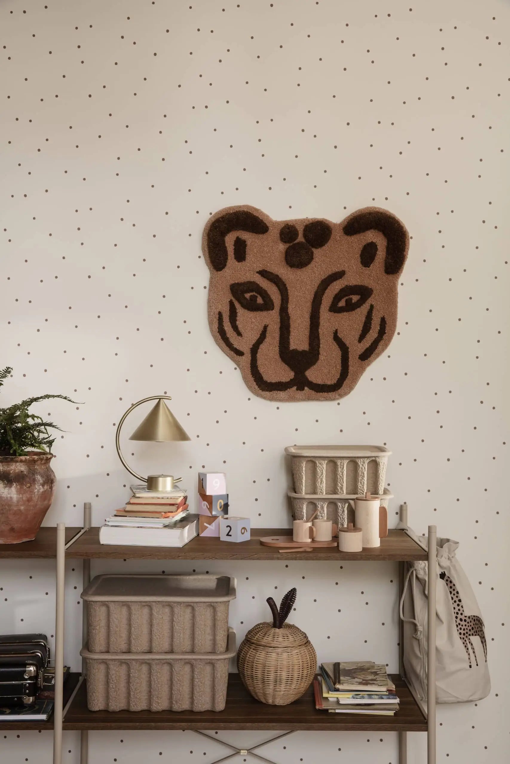 A cozy room features a wall with dot patterns, a leopard face rug, and a shelf with books, candles, baskets, and the sculptural ferm LIVING Meridian Lamp. A plant in a clay pot and a beige backpack hang on the right.