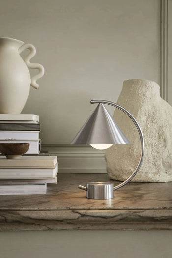 The ferm LIVING Meridian Lamp, featuring a conical shade and sculptural design, sits on a wooden surface beside stacked books and ceramic vases, adding minimalist style to the decor.
