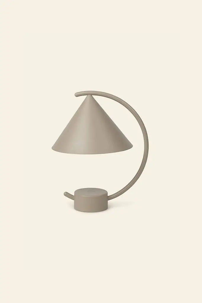The ferm LIVING Meridian Lamp by ferm LIVING is a modern minimalist table lamp with a sculptural form, conical shade, and curved arm in smooth beige powder-coated steel, shown against a plain light background.