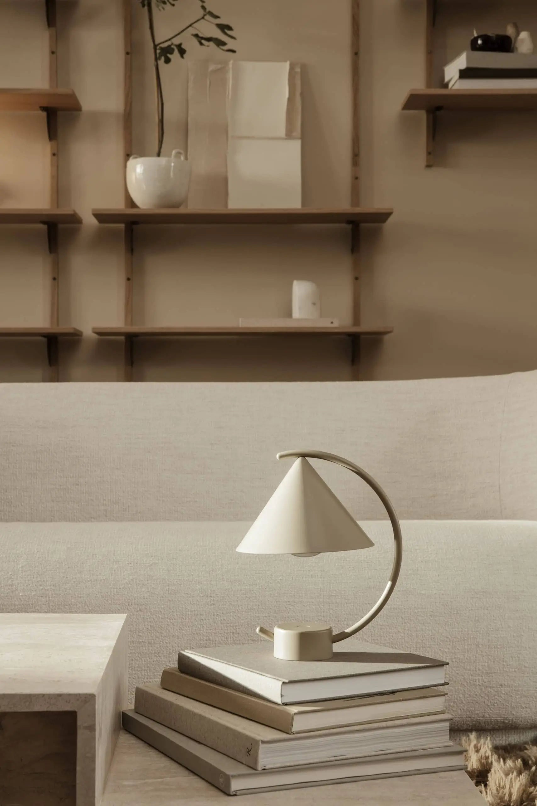 Minimalist living room with neutral tones, featuring the sculptural ferm LIVING Meridian Lamp on stacked books, a light sofa, and wall-mounted wooden shelves with understated decor in the background.