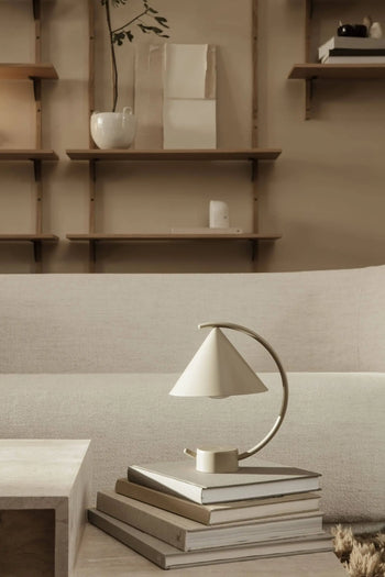 Minimalist living room with neutral tones, featuring the sculptural ferm LIVING Meridian Lamp on stacked books, a light sofa, and wall-mounted wooden shelves with understated decor in the background.