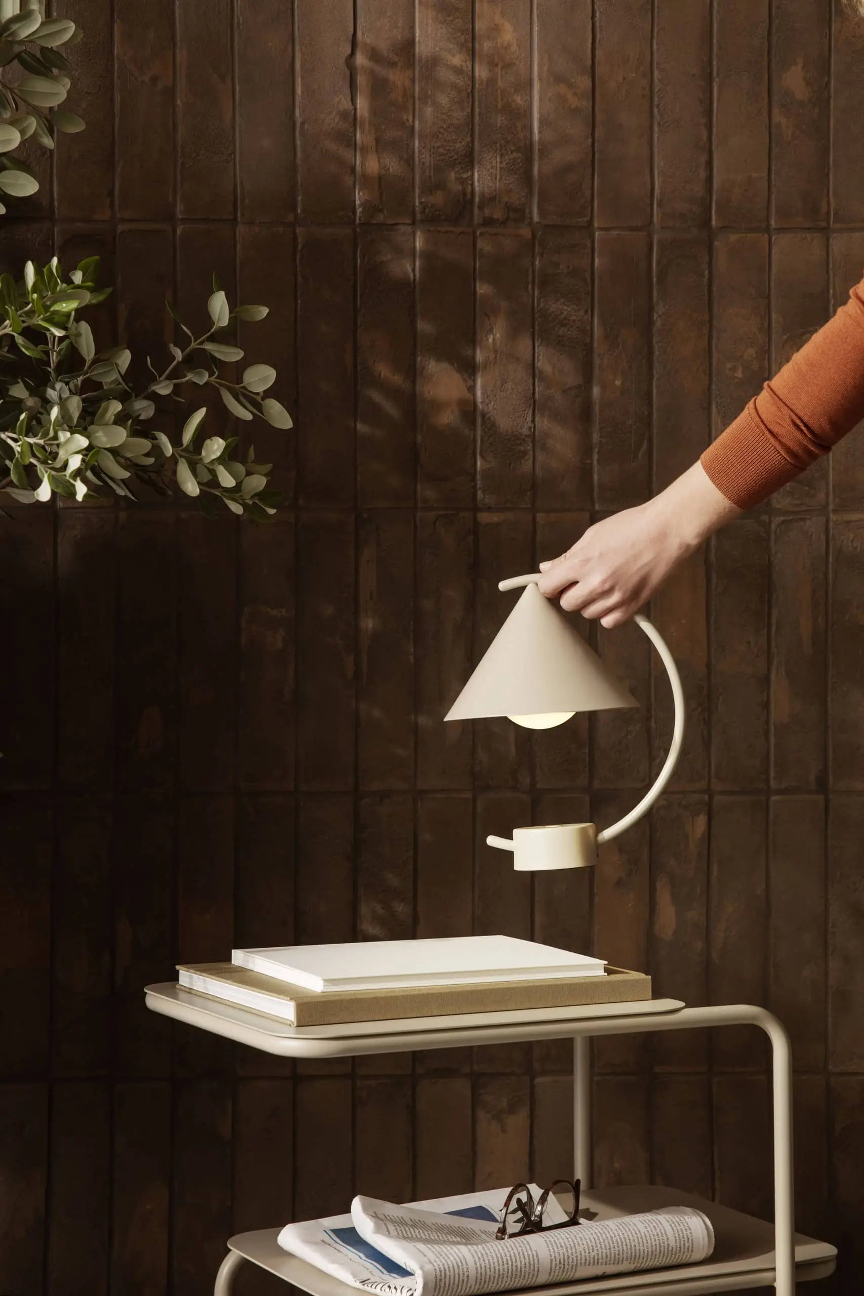 A hand adjusts the ferm LIVING Meridian Lamp—a modern white sculptural lamp—on a side table with books and glasses against a dark brown tiled wall. Green leaves appear on the left.