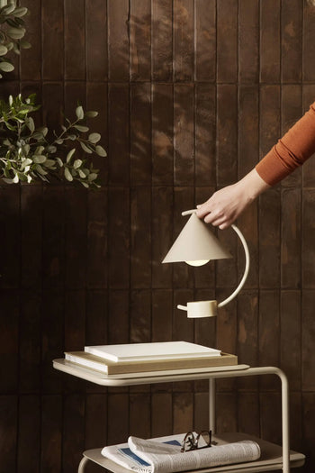 A hand adjusts the ferm LIVING Meridian Lamp—a modern white sculptural lamp—on a side table with books and glasses against a dark brown tiled wall. Green leaves appear on the left.