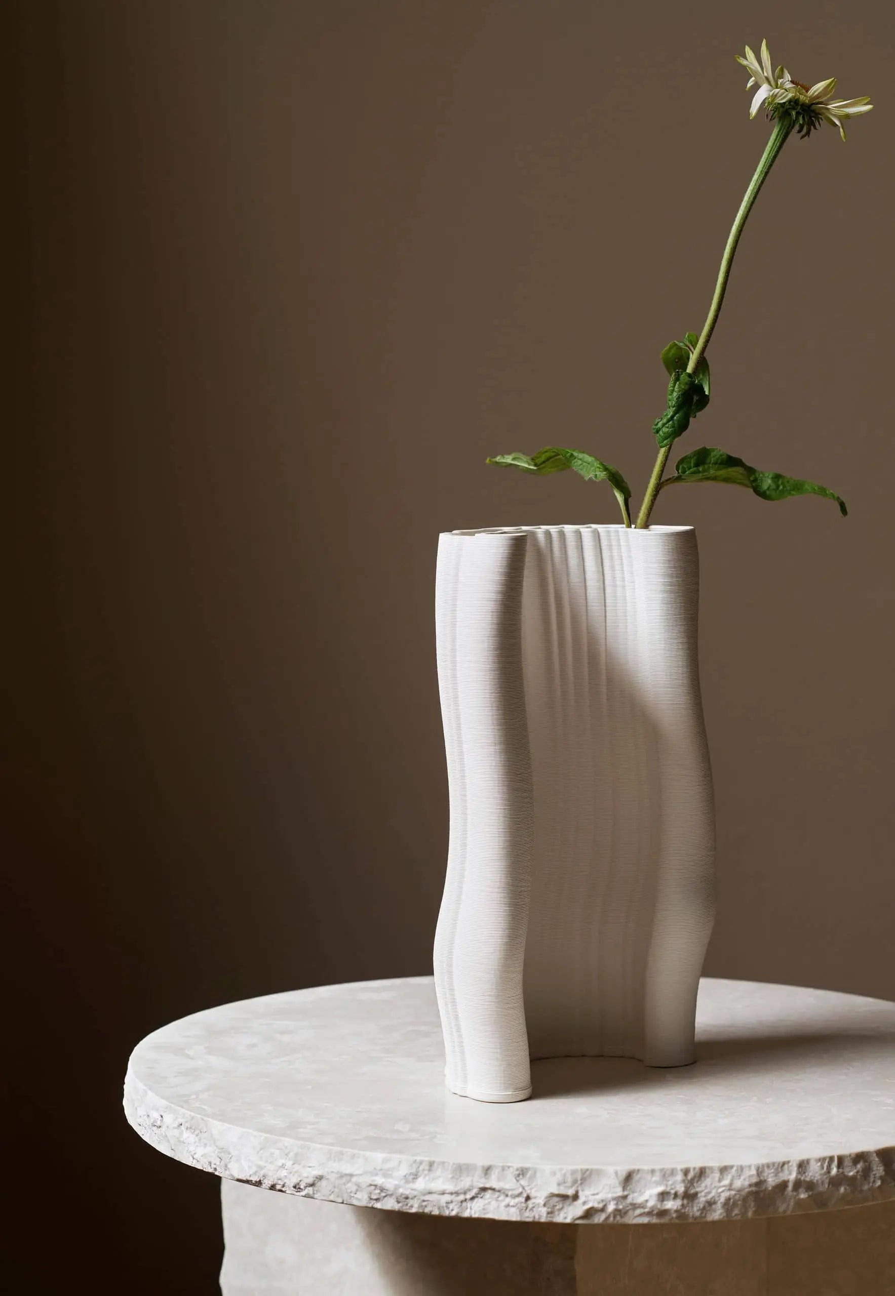 A single white flower with drooping petals and green leaves sits in the tall ferm LIVING Moire Vase on a round, light marble table against a neutral brown background.
