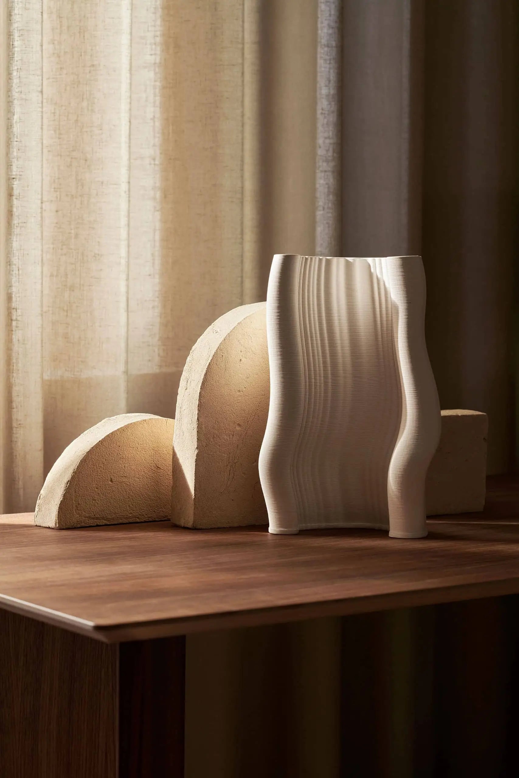 A wooden table displays three modern sculptures: two beige, semi-circular shapes and the ferm LIVING Moire Vase with wavy edges by ferm LIVING, crafted using 3D printing. Sheer curtains in soft light add an elegant charm.