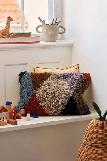 A colorful, textured pillow with geometric patterns rests on a white windowsill. Wooden toys, a giraffe figurine, and the ferm LIVING Mus Plant Pot, Large by ferm LIVING with mouse handles nearby create a cozy, playful atmosphere.