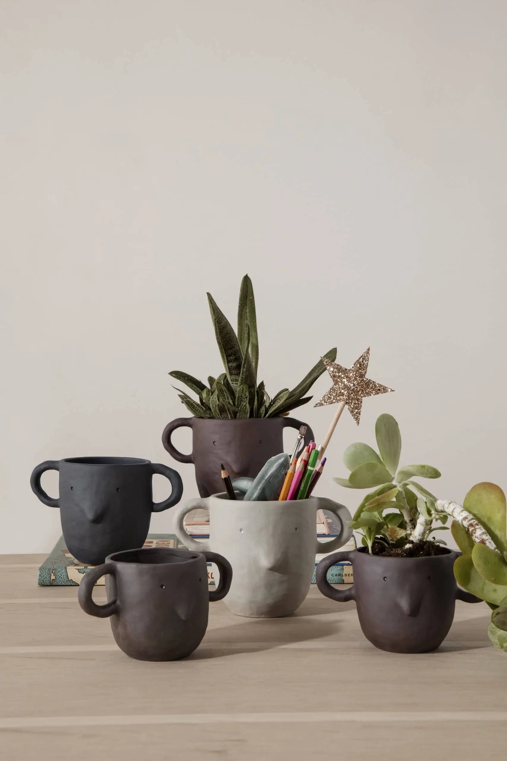 Five ferm LIVING Mus Plant Pots, Small—ceramic face-shaped mugs, some with mouse handles—sit on wood. Three display plants, one holds pens and a glittery star, and one is empty. Books and decor accent the background.