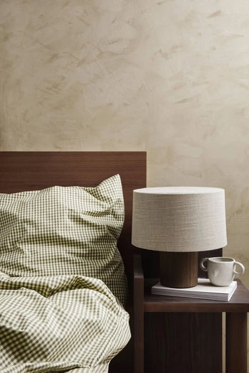 A neatly made bed with green and white checkered bedding is next to a wooden nightstand holding a ferm LIVING Mus Plant Pot, Small, a table lamp, a white cup, and two books against a beige textured wall.