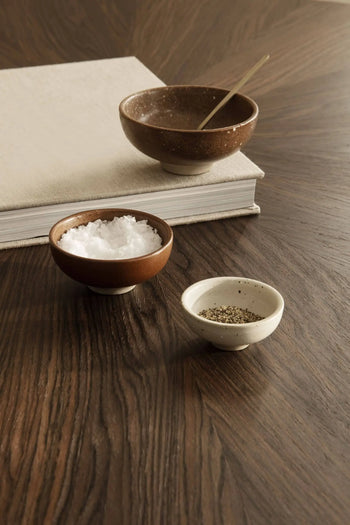 The ferm LIVING Petite Bowls, Set of 3, by ferm LIVING, are displayed on a wooden table holding salt, pepper, and a liquid with a spoon—ideal for adding elegance to your dining setup.