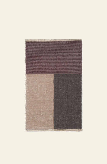 The ferm LIVING Pile Bathroom Mat features ribbed dark maroon, smooth beige (left), and dark brown (right) color blocks. Made from recycled polyester for a luxurious feel, it’s set against a light beige background.