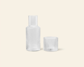 The ferm LIVING Ripple Carafe Set, Small features a clear, ribbed carafe and matching cup crafted from mouth-blown glass, both displayed against a plain, light beige background.