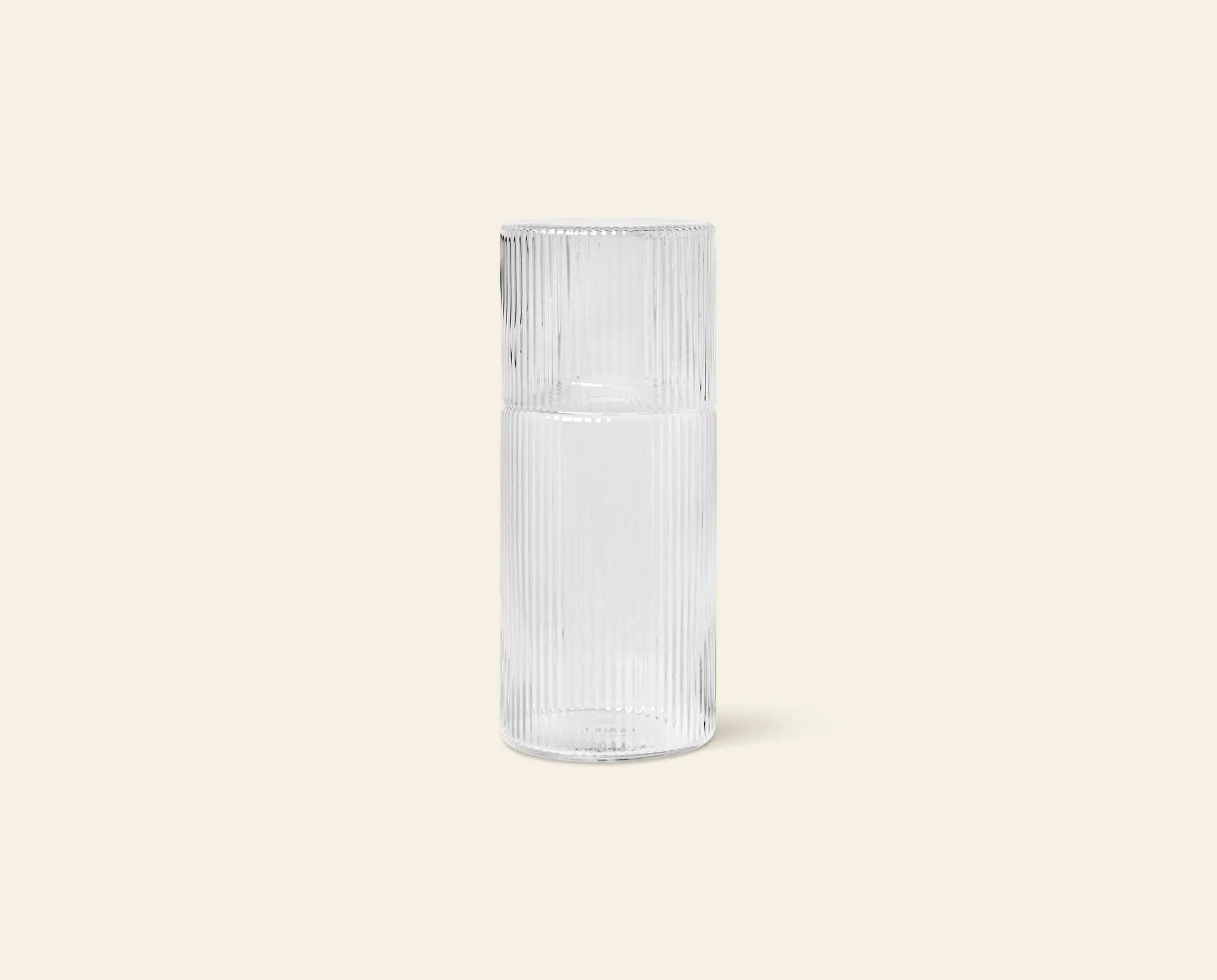 The ferm LIVING Ripple Carafe Set, Small, features a clear, tall cylindrical carafe with a ribbed texture and narrow neck. Crafted from mouth-blown glass, the inverted matching cup serves as a stylish lid. Shown on a plain beige background.
