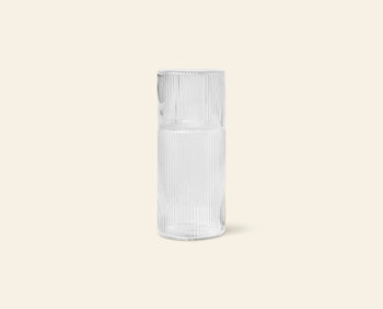 The ferm LIVING Ripple Carafe Set, Small, features a clear, tall cylindrical carafe with a ribbed texture and narrow neck. Crafted from mouth-blown glass, the inverted matching cup serves as a stylish lid. Shown on a plain beige background.